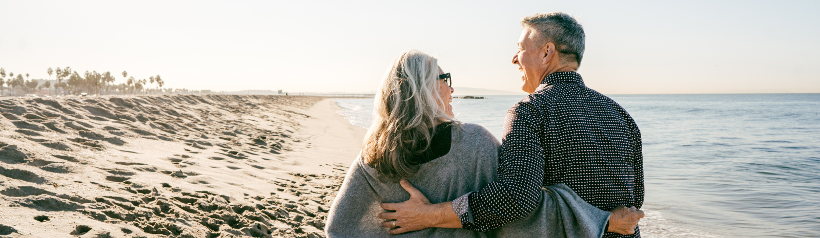 Older couple walking on beach with arms around each others backs.