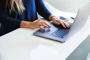 Close up of woman in blue shirt in front of white desk with both hands on her laptop.
