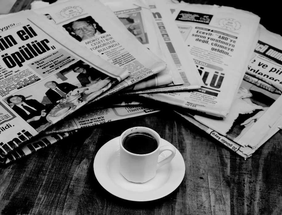 Black and white photo of a stack of newspapers spread around a cup of coffee.