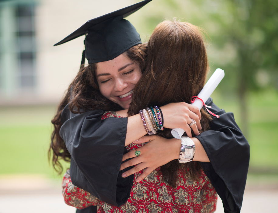 Young lady graduate and mother sharing a hug as she graduates as she is in her graduation gown and holding a diploma.