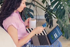 Woman in pink shirt sitting on a couch, holding a cup of coffee and a debit card with a laptop on the sign in screen of online banking.