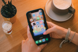Close up of woman's hands holding her phone looking at her apps with a coffee, candle, and plant on a wooden table.