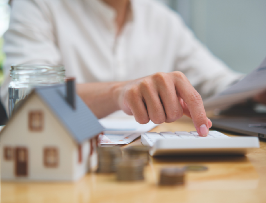 Woman in white shirt at desk in front of laptop, holding paper while punch numbers into calculator, with miniature house and coins on the desktop.